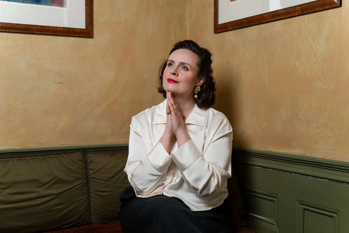 Katy Thomson seated in a white blouse, looking upwards with her hands together